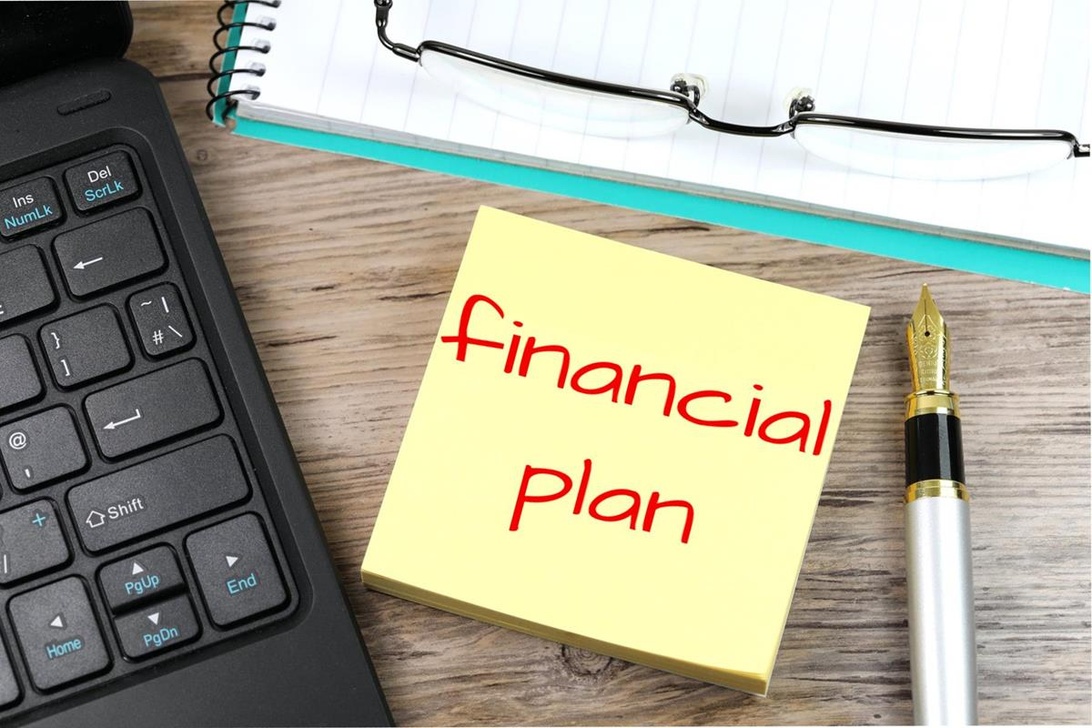 En arbetsplats med en gul kom-ihåg-lapp med texten 'Financial Plan', laptop, anteckningsbok och penna, vilket representerar processen för finansiell planering.